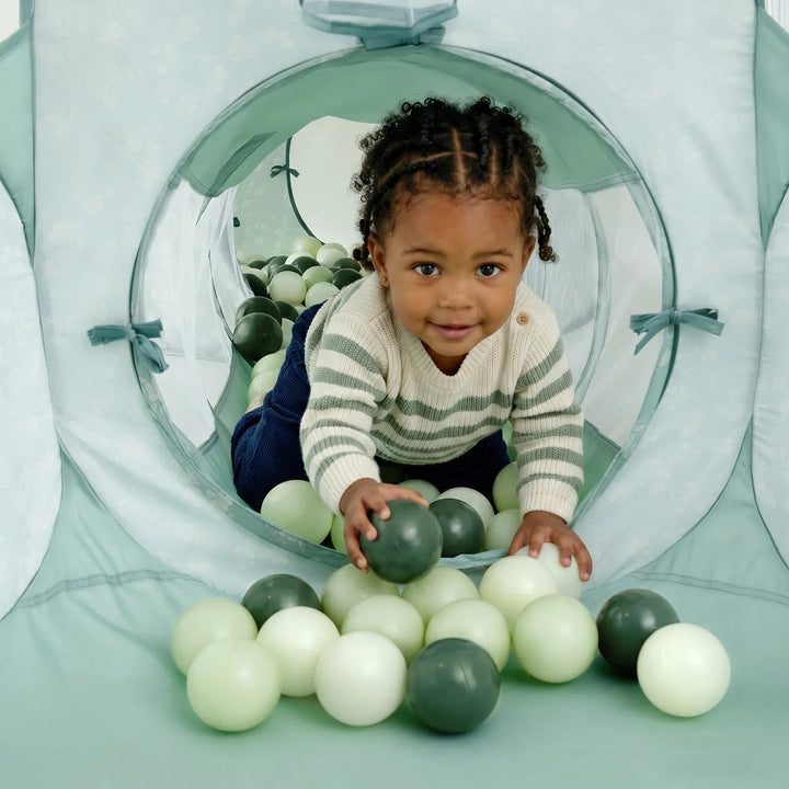 A toddler crawling through a Sea Dragon Dreams 3-in-1 play tunnel filled with sage green and white plastic balls.