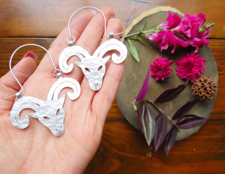 Sea Dragon Dreams handmade aluminum Aries ram hoop earrings with hammered texture held in a hand near pink flowers.