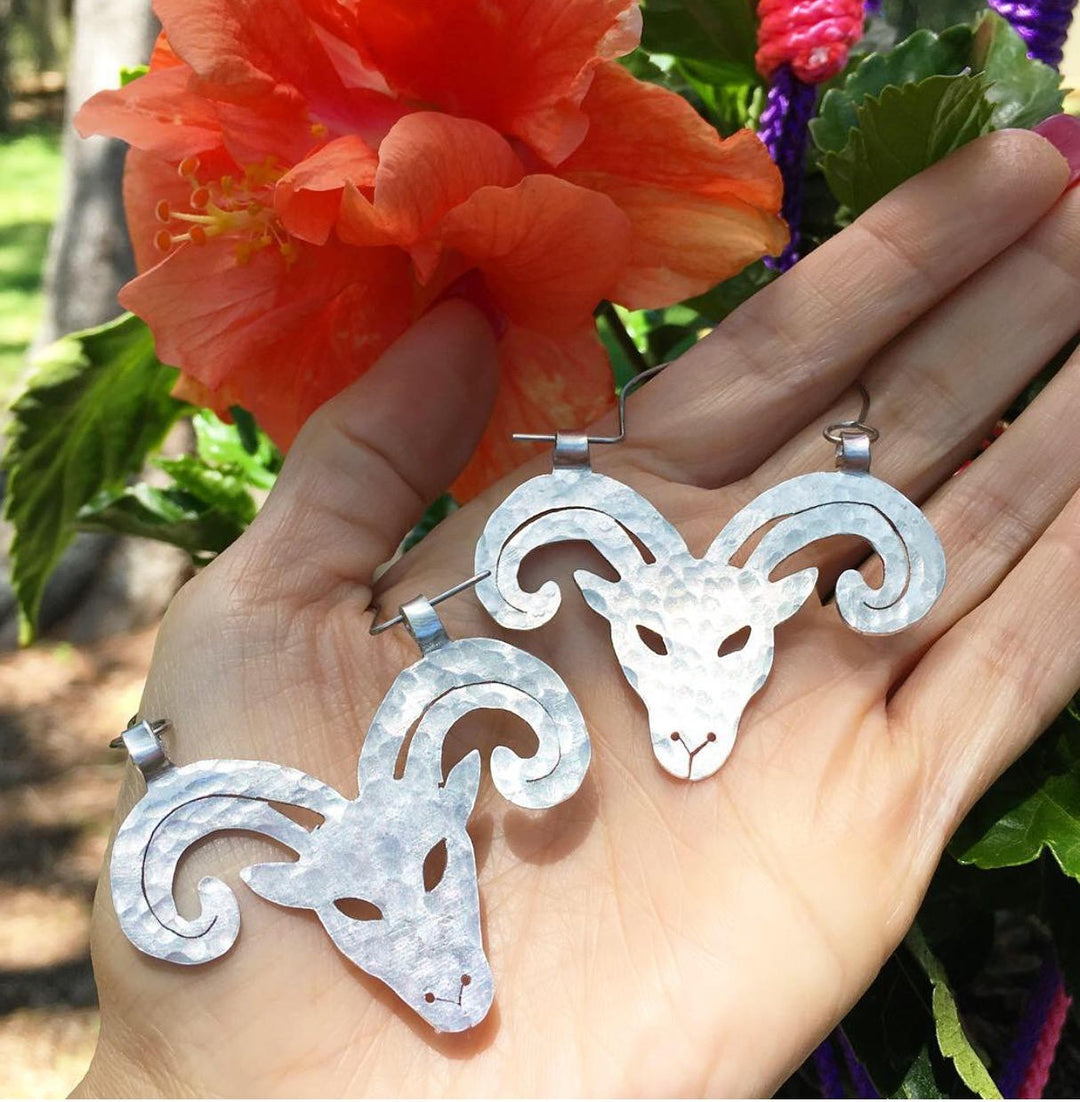 Sea Dragon Dreams handmade aluminum Aries ram hoop earrings with a hammered texture held in a hand near a hibiscus.