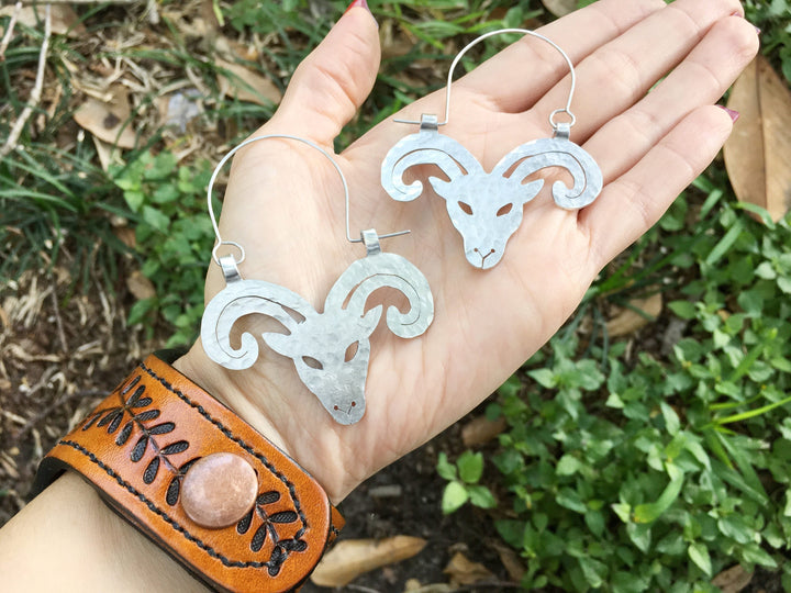 Sea Dragon Dreams handmade aluminum Aries ram hoop earrings with a hammered texture held in a palm over greenery.