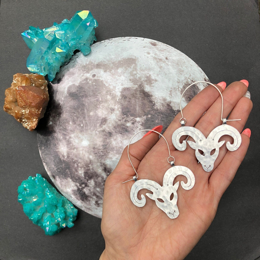 Sea Dragon Dreams handmade aluminum Aries ram hoop earrings held in a hand over a moon backdrop with crystals.