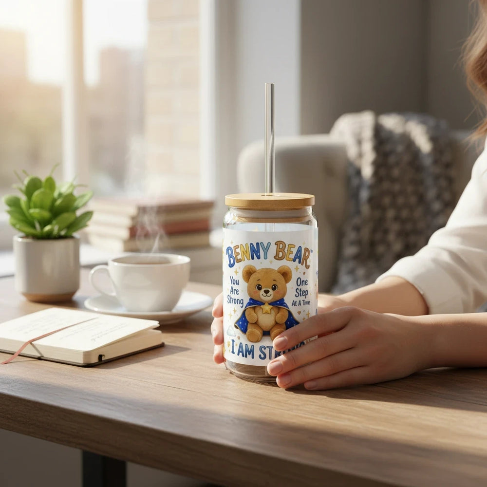 Sea Dragon Dreams Benny Bear I Am Strong 16oz glass sipper with bamboo lid and straw held by hands on a wooden desk