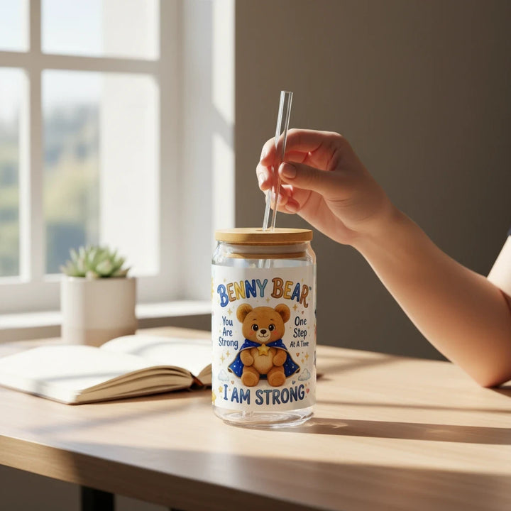 Sea Dragon Dreams Benny Bear I Am Strong 16oz glass sipper with bamboo lid and straw held by a hand on a desk.