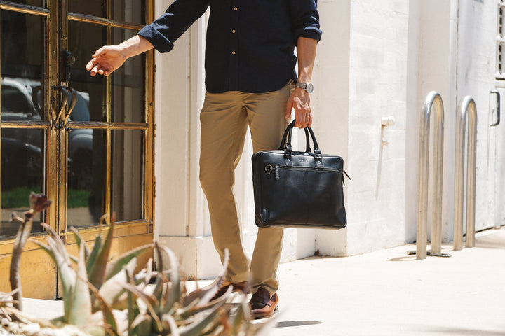 A man in business casual attire carrying a black Sea Dragon Dreams slim leather laptop bag outside an office building.