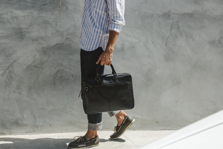 Man carrying a black Sea Dragon Dreams slim leather laptop bag with nickel hardware while walking outdoors.