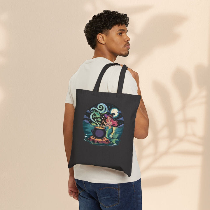 Sea Dragon Dreams black cotton canvas tote bag featuring a whimsical mermaid cauldron design worn over the shoulder.