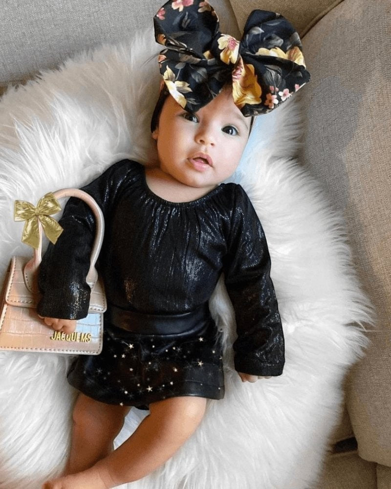 Baby wearing a Sea Dragon Dreams onyx black shimmer long sleeve leotard with a floral bow headband on a white rug