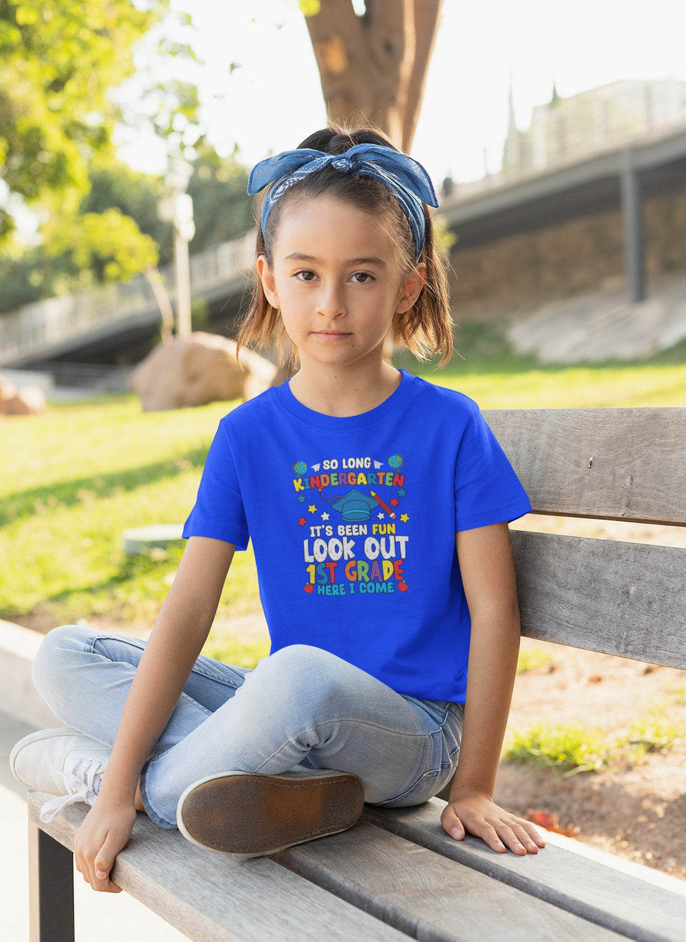 Sea Dragon Dreams blue youth t-shirt with Look Out First Grade graphic worn by a girl sitting on a wooden bench.