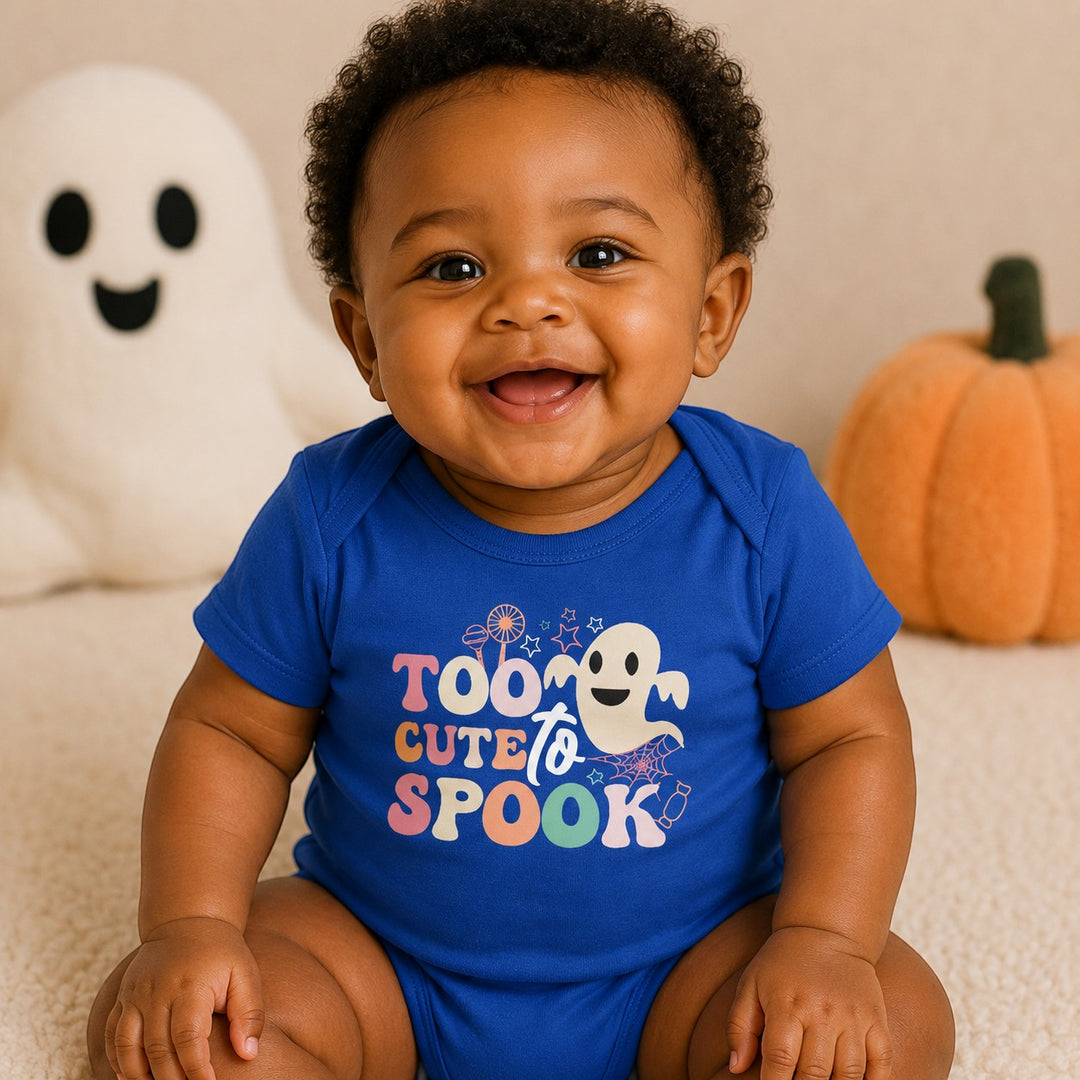A smiling baby wearing a blue Sea Dragon Dreams bodysuit with a Too Cute To Spook Halloween graphic.