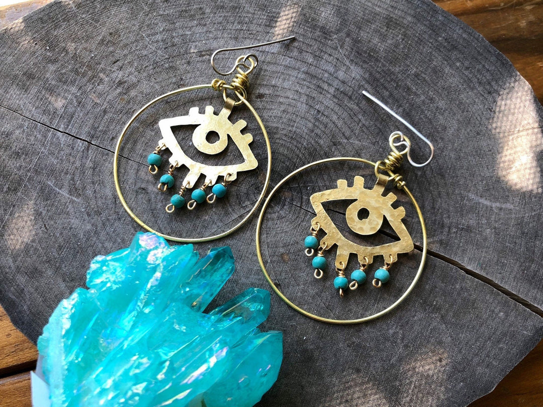 Sea Dragon Dreams handmade brass evil eye hoop earrings with blue magnesite beads on a rustic wood surface.