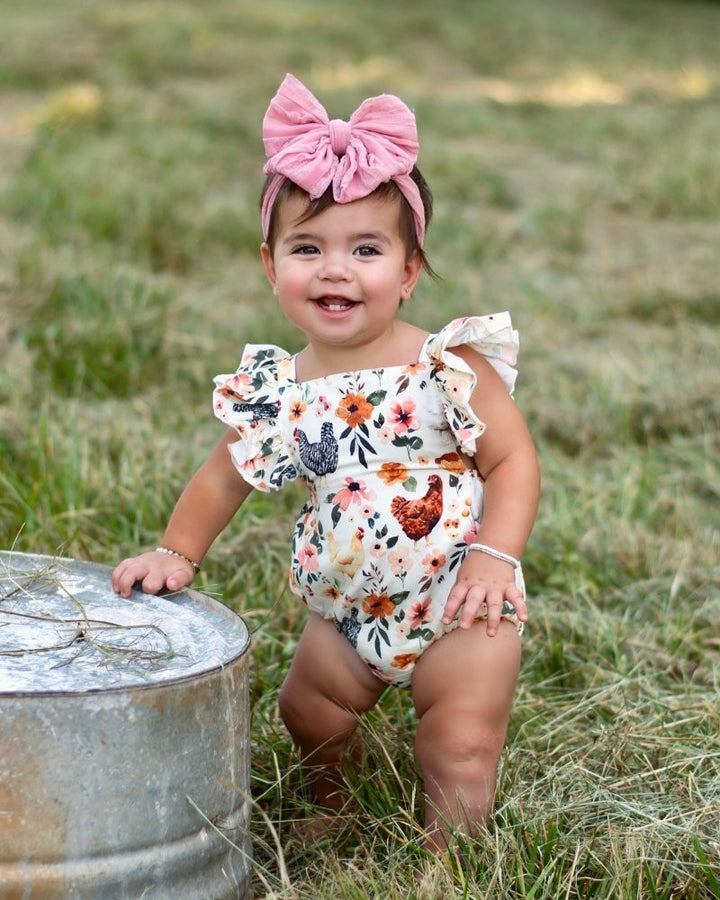 Sea Dragon Dreams baby girl wearing a cream romper with a chicken and floral print and ruffle shoulder straps