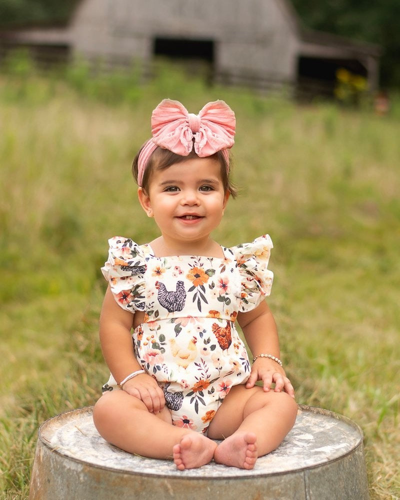 Sea Dragon Dreams baby girl wearing a white romper with a chicken print and ruffle straps, sitting on a metal tub.
