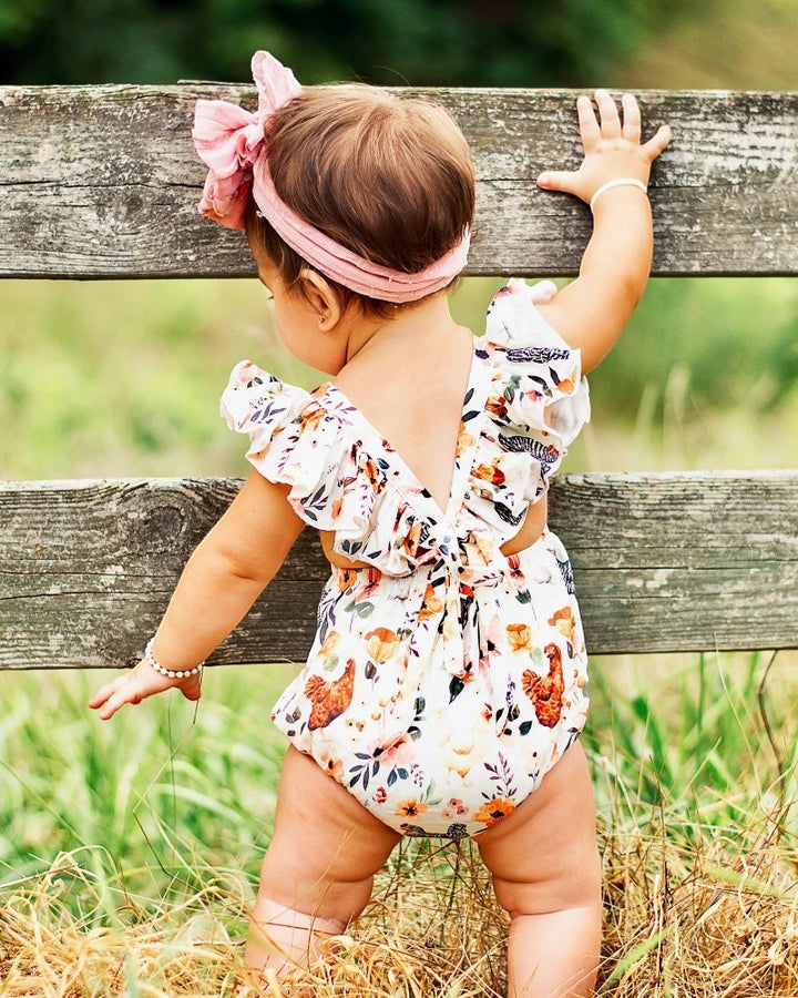 Sea Dragon Dreams baby romper with chicken print, featuring ruffle straps and a tie-back detail, worn outdoors.