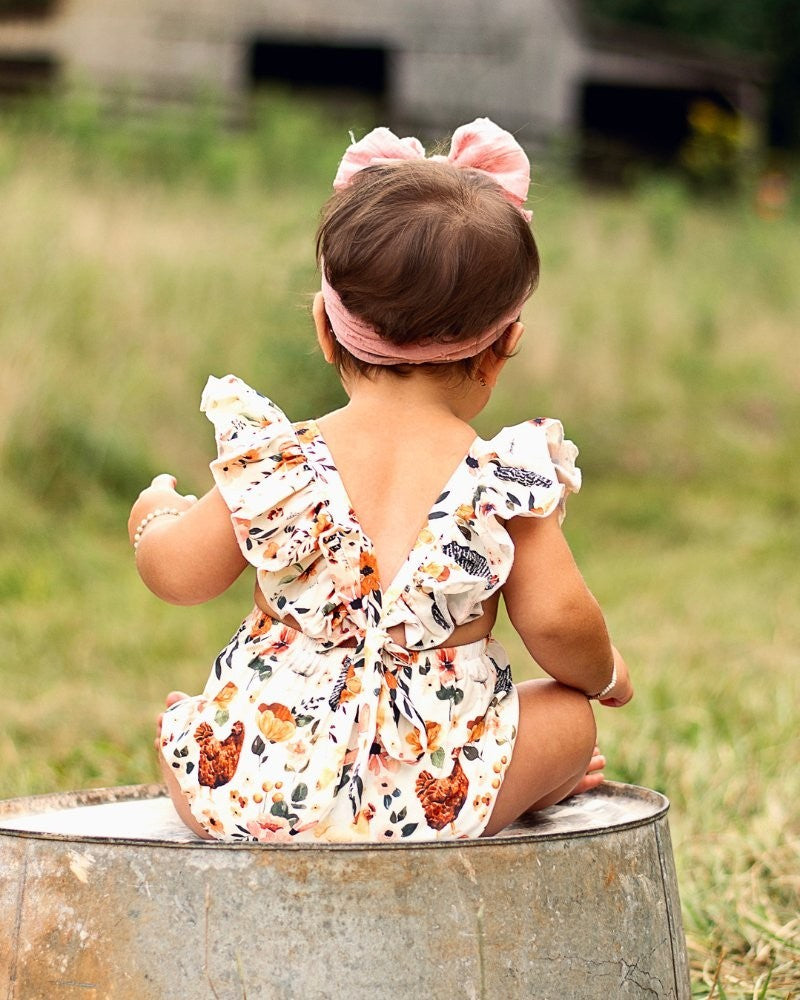 Sea Dragon Dreams baby romper with chicken print, ruffle back straps, and bow detail, worn by a toddler outdoors.