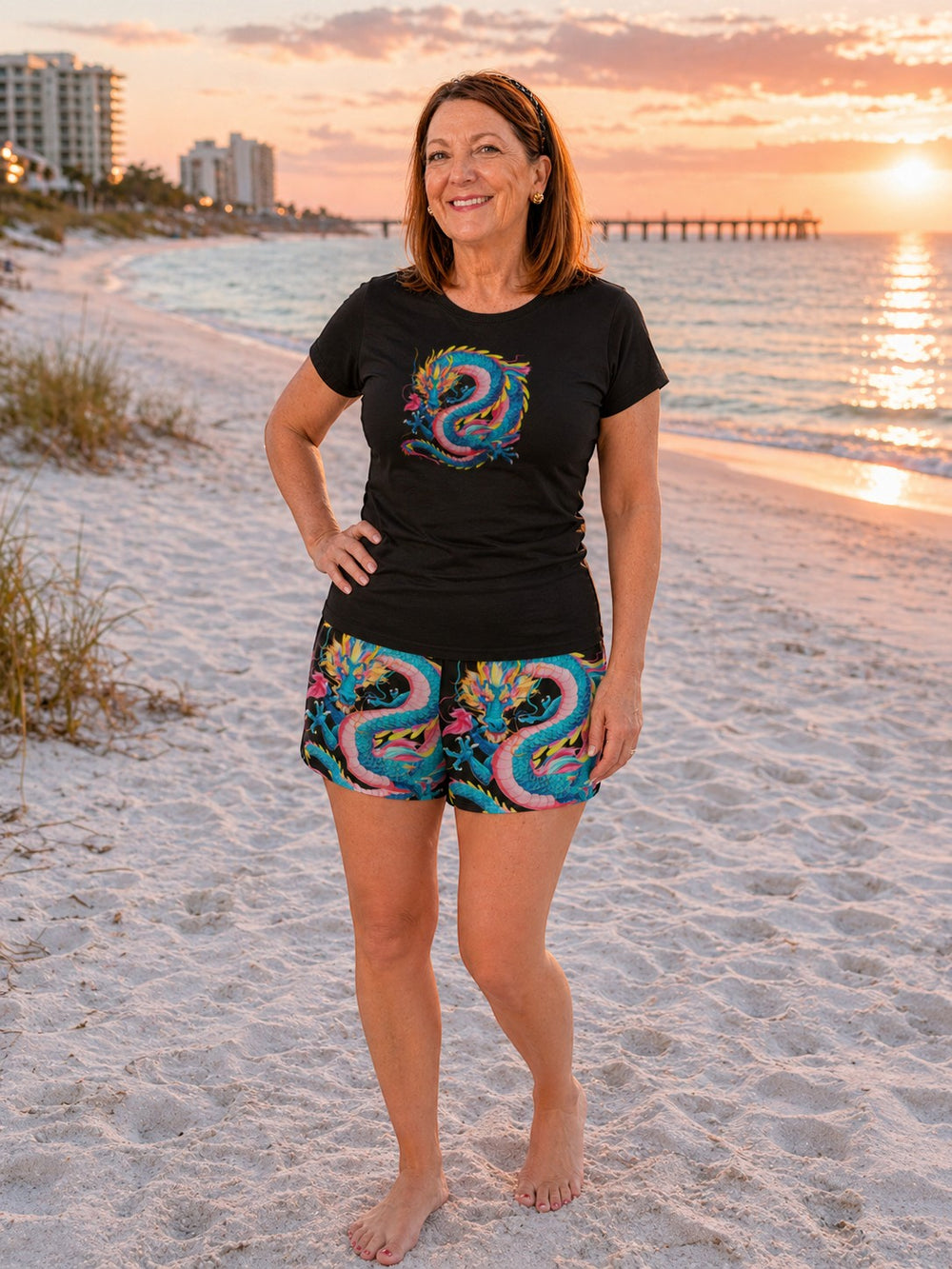 A woman wearing Sea Dragon Dreams shorts with a vibrant dragon print on a sandy beach at sunset.