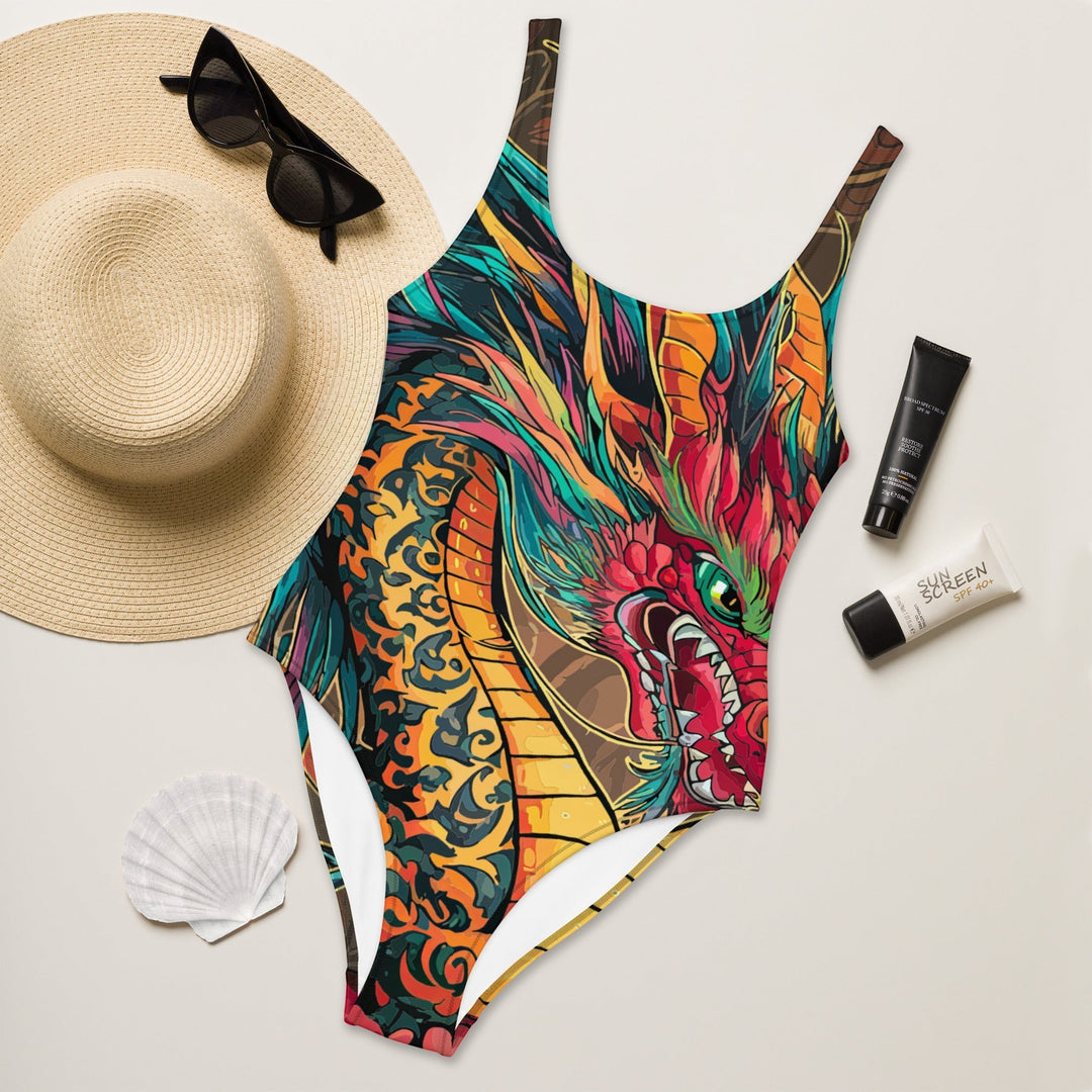 Sea Dragon Dreams one-piece swimsuit with a vibrant dragon print, scoop neck, and cheeky fit on a beach-themed flat lay.