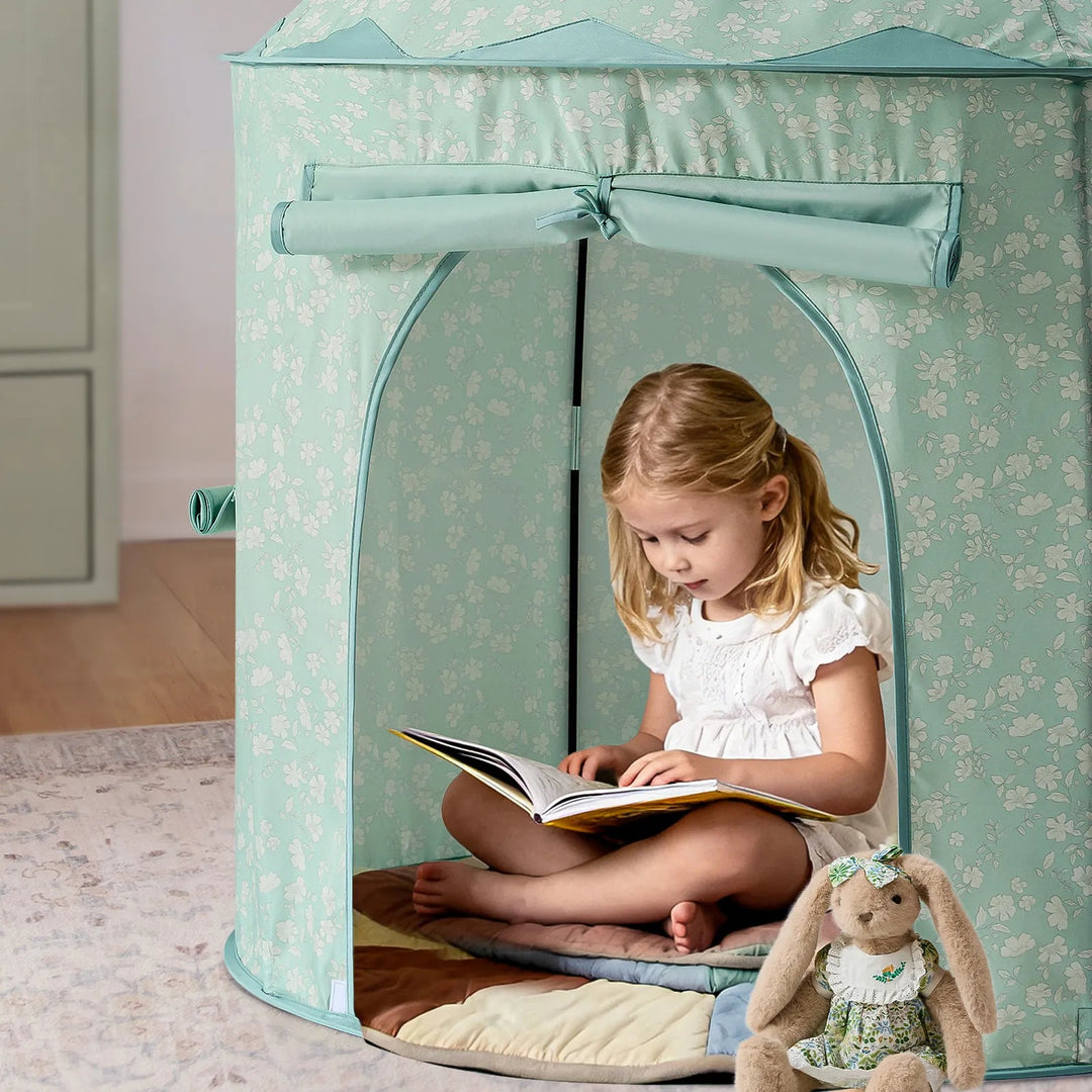 A young girl reading inside a Sea Dragon Dreams mint green floral play tent with a plush toy sitting nearby.