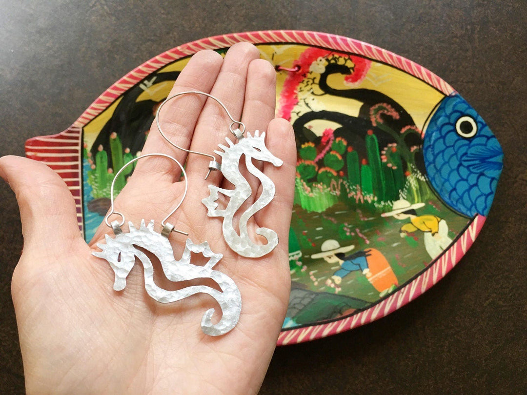 Sea Dragon Dreams handmade silver-tone hammered metal seahorse hoop earrings held in a hand over a painted fish dish.