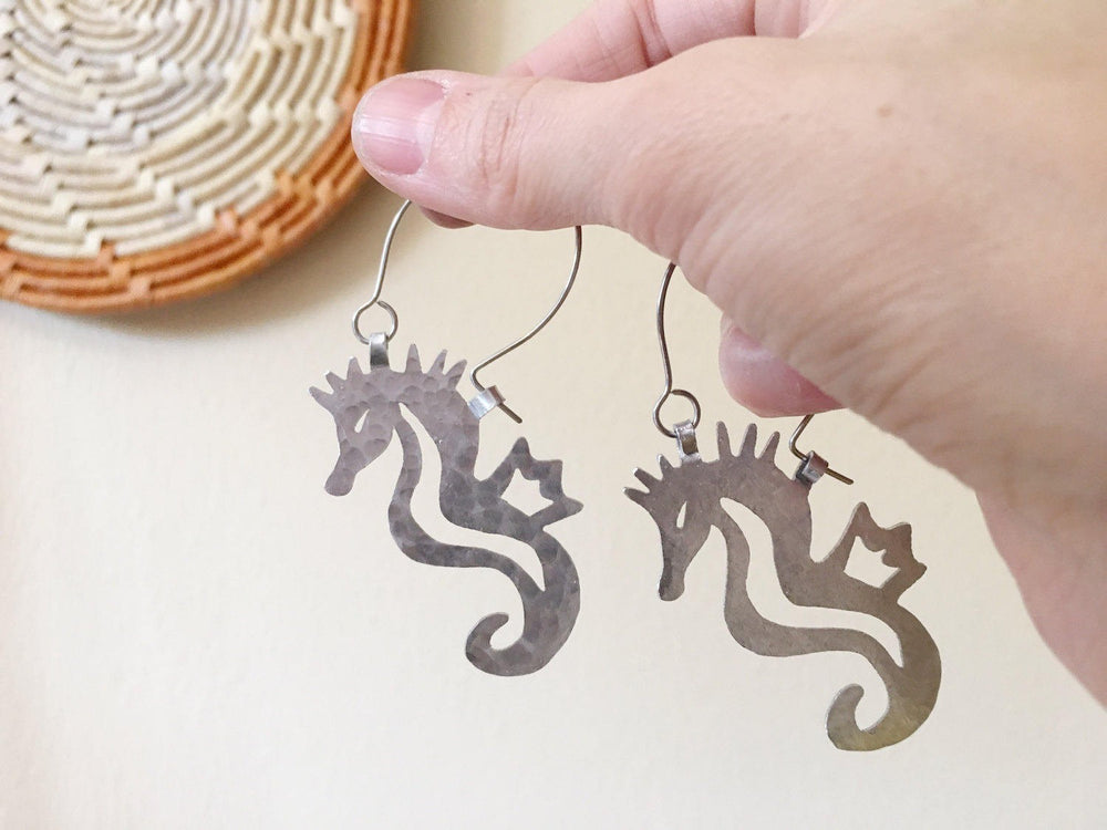Sea Dragon Dreams handmade seahorse hoop earrings with a hammered silver-tone finish held in a hand.
