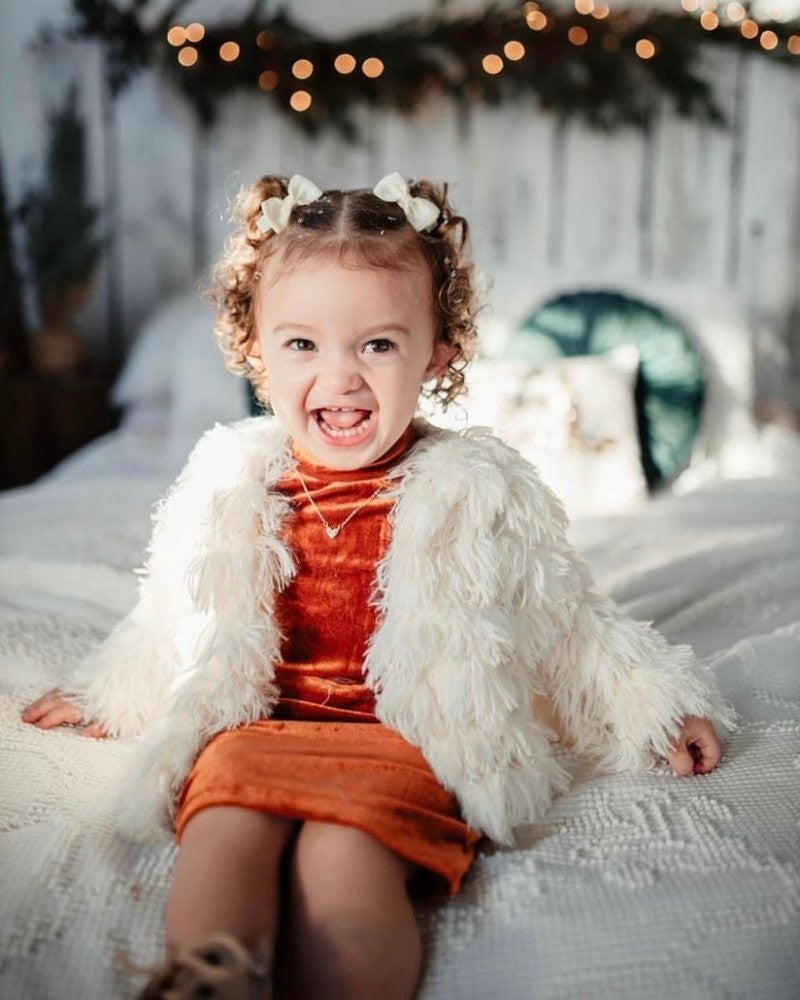 Sea Dragon Dreams ivory faux shag layered jacket worn by a smiling toddler girl sitting on a white bed