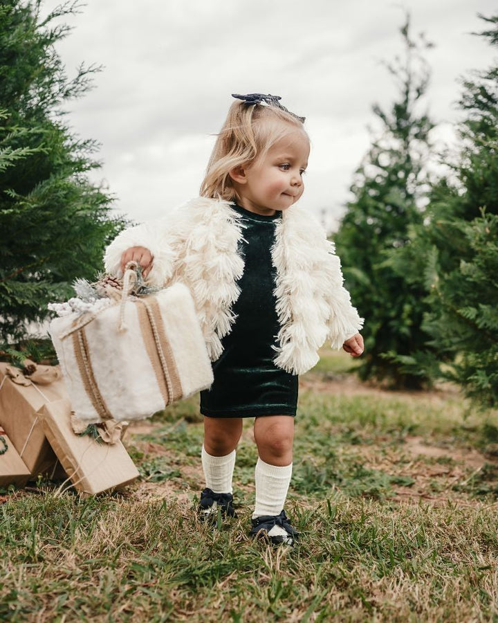 Toddler girl wearing a Sea Dragon Dreams ivory faux shag layered jacket over a dark green dress in a pine forest.