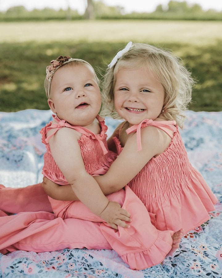 Two young girls wearing Sea Dragon Dreams Maisie smocked two-piece sets in pressed peony pink on a floral blanket.