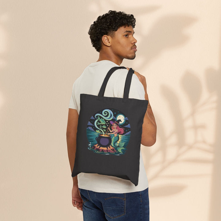 Sea Dragon Dreams black cotton canvas tote bag featuring a whimsical mermaid cauldron design worn over the shoulder.