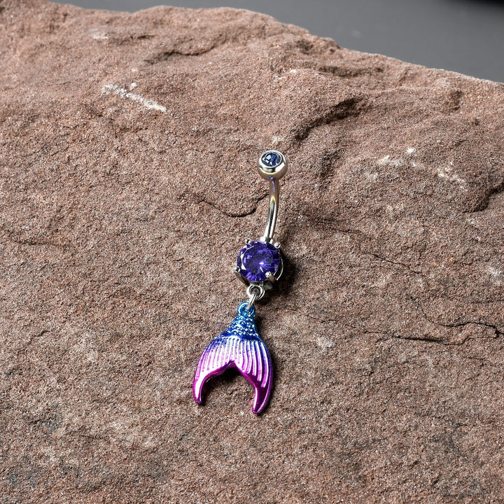 Sea Dragon Dreams purple gemstone belly ring featuring an aqua and pink enamel mermaid tail dangle on a stone surface
