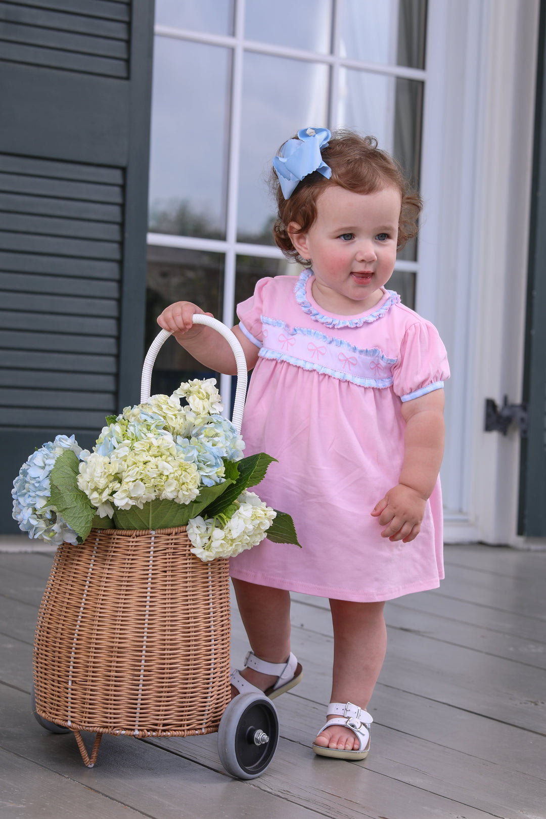 A toddler girl wearing a Sea Dragon Dreams pink dress with puff sleeves and bow detail, standing with a flower basket.