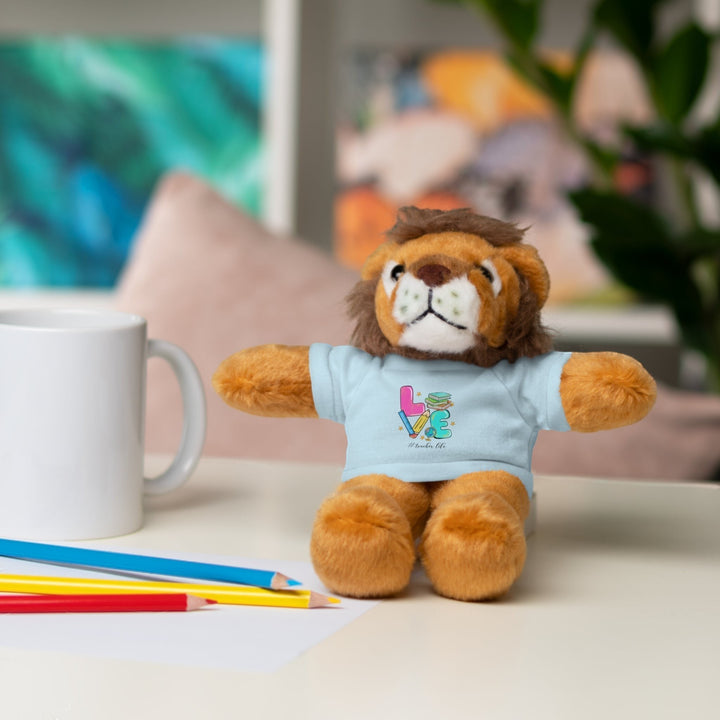 Sea Dragon Dreams plush lion stuffed animal wearing a light blue t-shirt with Love Teacher Life graphic on a desk.