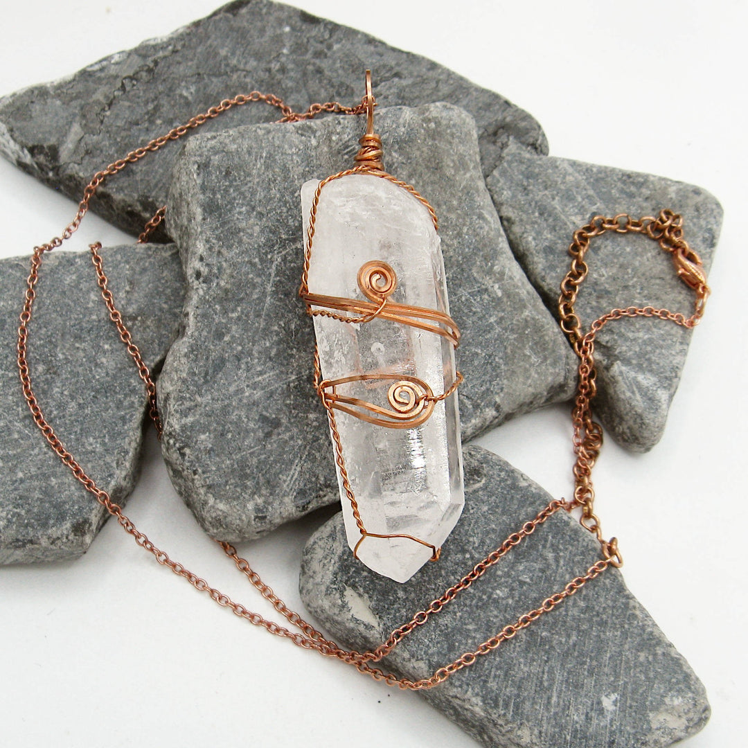 Sea Dragon Dreams raw clear quartz crystal pendant wire-wrapped in solid copper on a matching chain over grey stones.