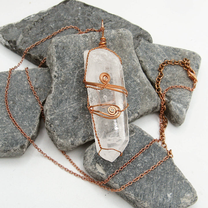 Sea Dragon Dreams raw clear quartz crystal pendant wire-wrapped in solid copper on a matching chain over grey stones.