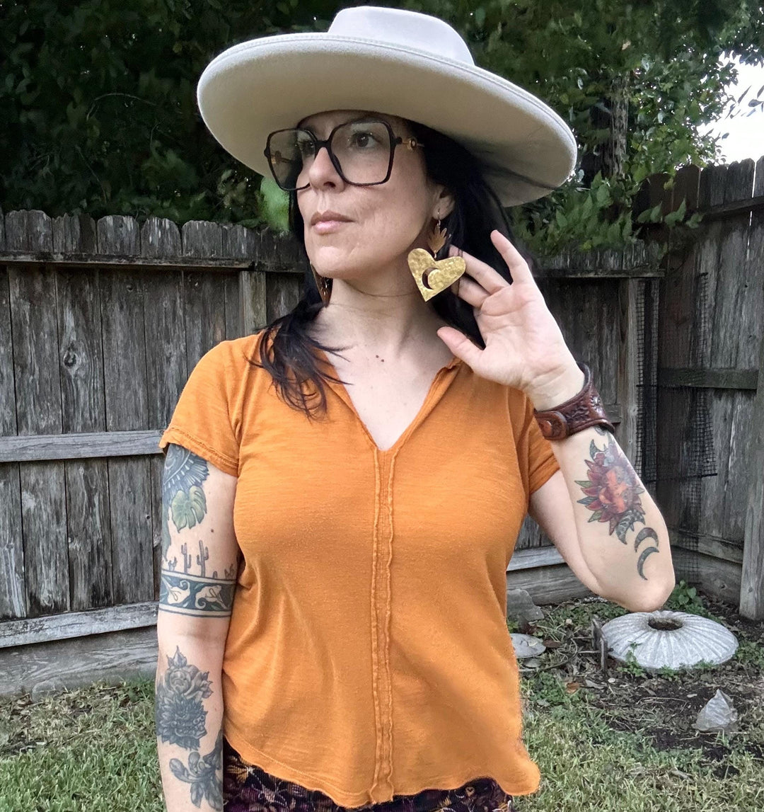 Sea Dragon Dreams XL sacred heart brass earrings with sun and moon design worn by a woman in a hat and orange shirt