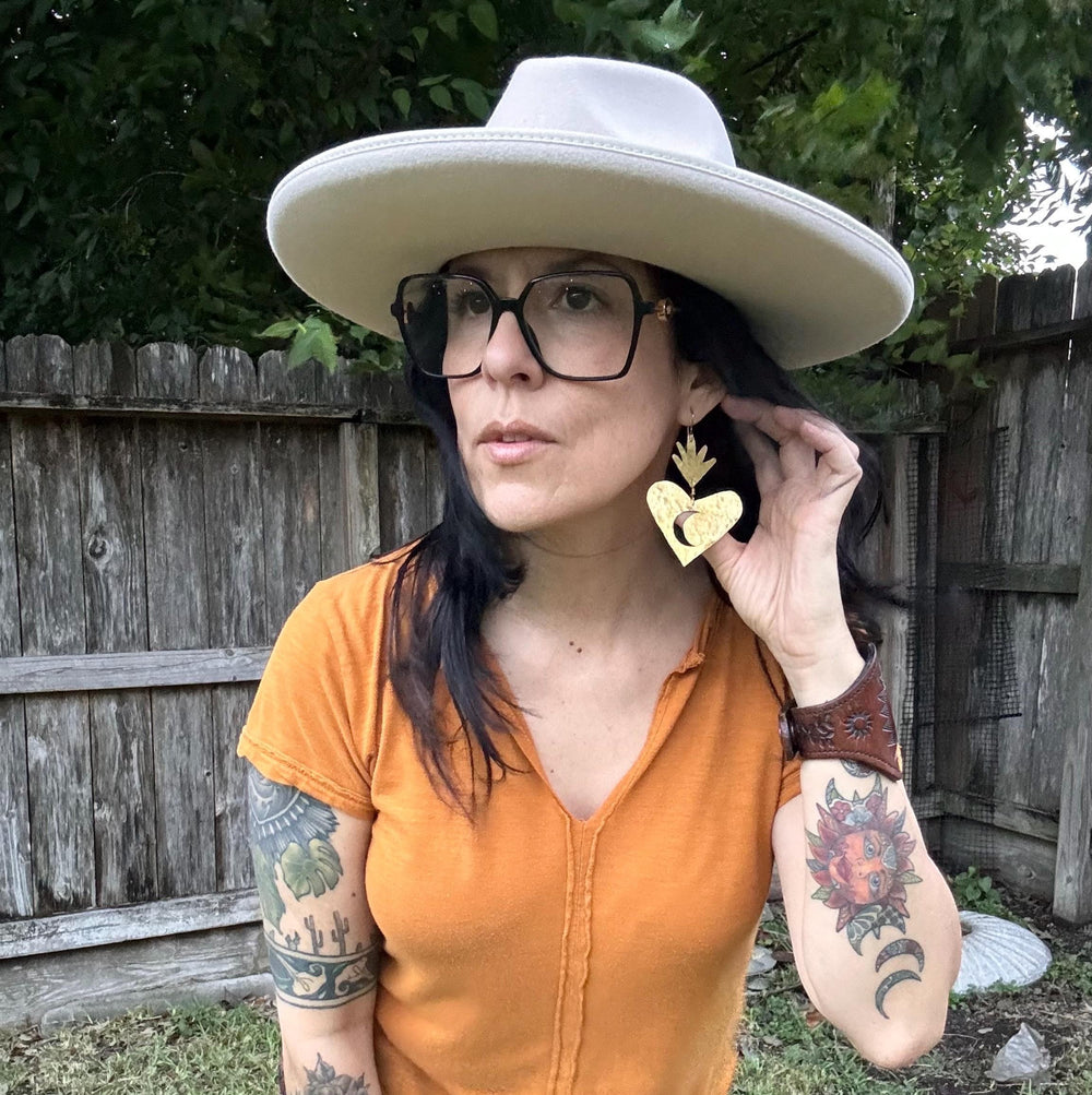 Sea Dragon Dreams XL sacred heart brass earrings with sun and moon cutouts worn by a woman in a wide-brimmed hat.