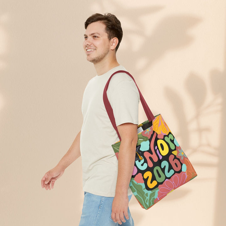 A man wearing a neutral t-shirt carries a Sea Dragon Dreams Senior 2026 tote bag with a colorful floral graphic.