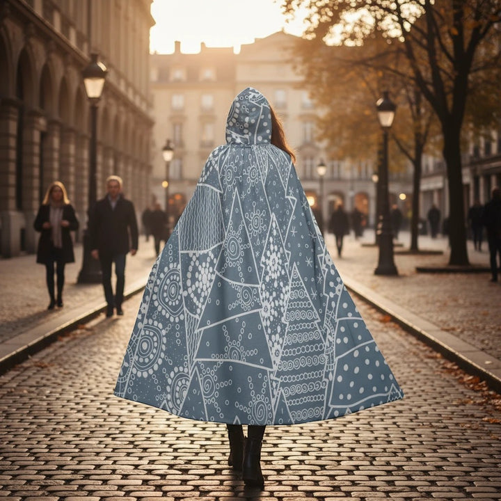 Sea Dragon Dreams unisex hooded cloak in blue with white geometric snowflake patterns, worn on a cobblestone street.