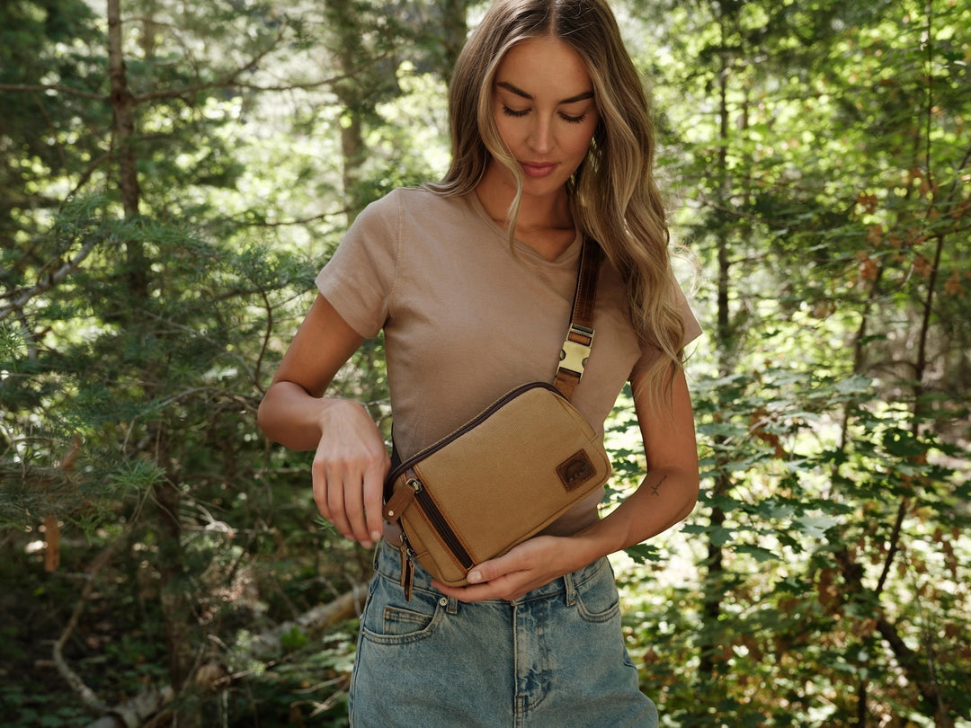 A woman wearing a Sea Dragon Dreams tan waxed canvas sling bag with brown leather accents in a forest setting.