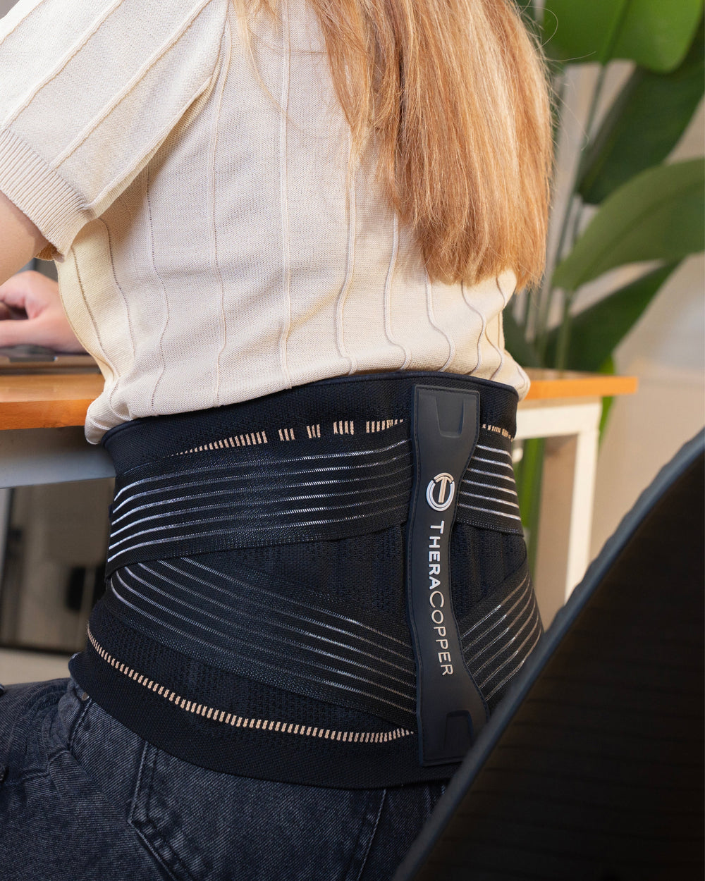 Sea Dragon Dreams TheraCopper back brace worn by a woman sitting at a desk, featuring black mesh and support bars.
