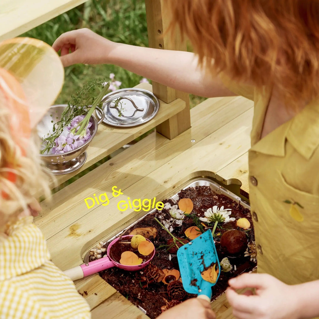 Sea Dragon Dreams Tiny Land mud kitchen with two children playing with soil, flowers, and tools in a wooden basin