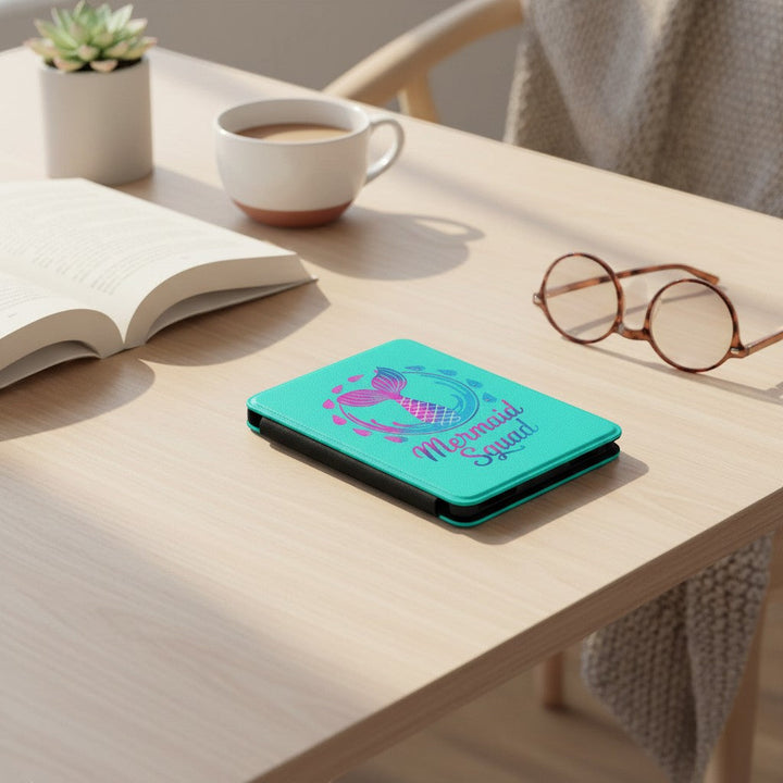 Sea Dragon Dreams turquoise Kindle case with Mermaid Squad tail design, resting on a wooden desk with a book and glasses.