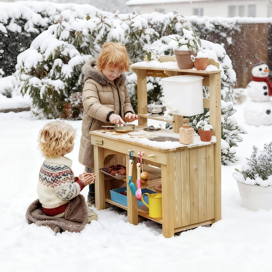Two children playing with a Sea Dragon Dreams Tiny Land wooden mud kitchen in a snowy backyard setting.