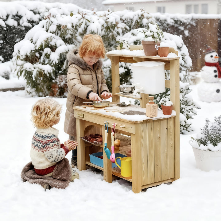 Two children playing with a Sea Dragon Dreams Tiny Land wooden mud kitchen in a snowy backyard setting.