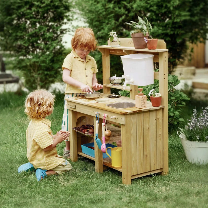 Two children playing with a Sea Dragon Dreams Tiny Land wooden mud kitchen in a lush green backyard garden.