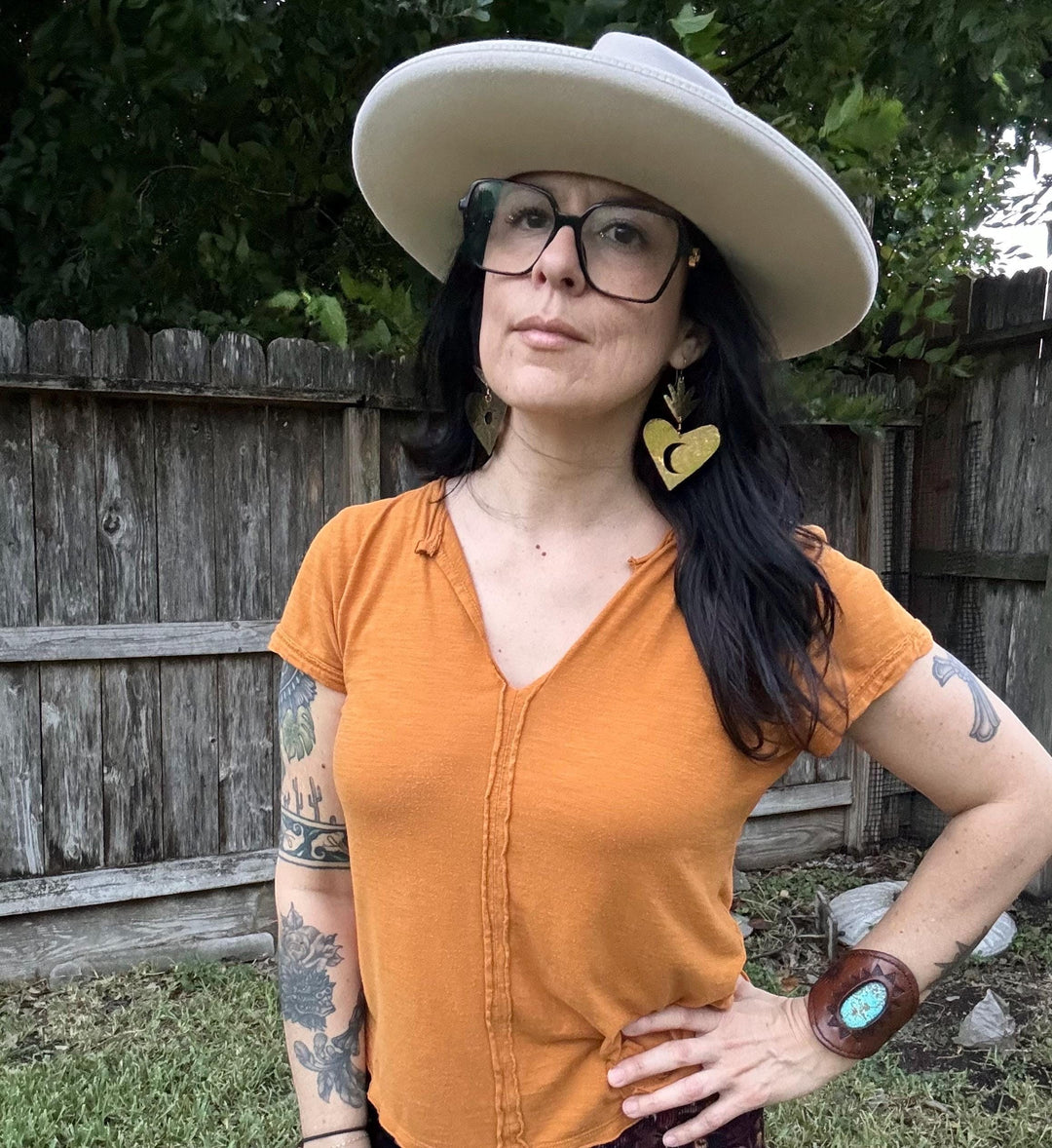 Sea Dragon Dreams XL brass sacred heart earrings with moon design worn by a woman in a wide-brim hat and orange top.