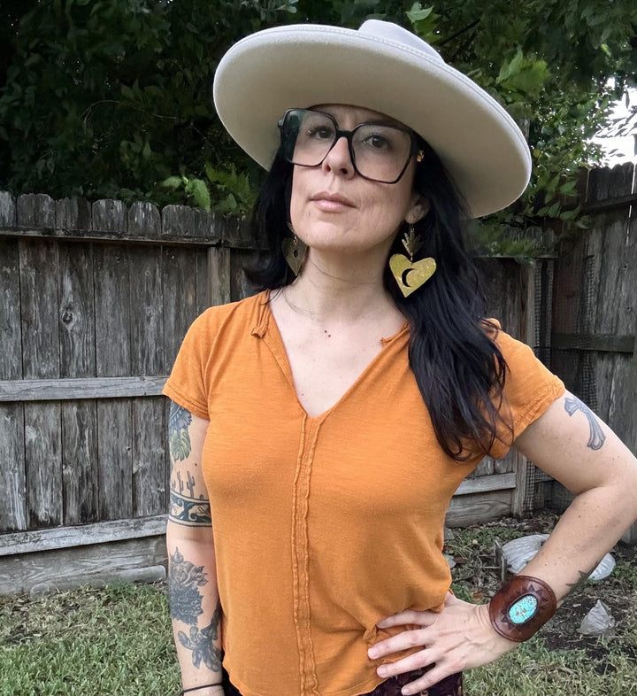 Sea Dragon Dreams XL brass sacred heart earrings with moon design worn by a woman in a wide-brim hat and orange top.